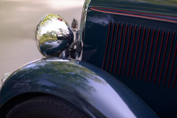 Chrome bullet headlamp, red-pinstriped louvers and glossy green fender of a vintage roadster gleam in sunlight on a park lane, showcasing elegant pre-war automotive design.