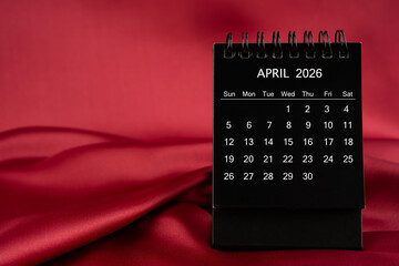 Black desk calendar for April 2026 on a red color satin or silk textile background.
