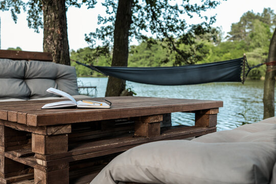 Notebook with pencil on rustic wooden pallet table, surrounded by soft cushions and a hammock by a peaceful lakeside, ideal for writing and relaxing in nature - Powered by Adobe