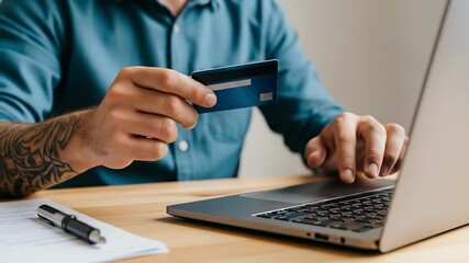 A man with a tattooed arm uses a blue credit card to make an online purchase on his laptop at a wooden desk.