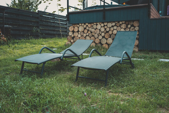 Two modern outdoor lounge chairs placed on green grass beside a cabin deck with stacked firewood. Cozy spot for summer sunbathing and countryside relaxation