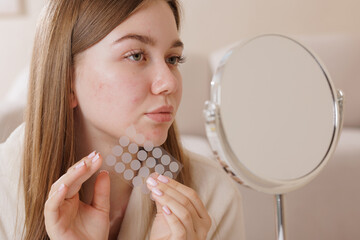 Teenager with skin blemishes holds acne patches by her face in front of a mirror, showing modern...