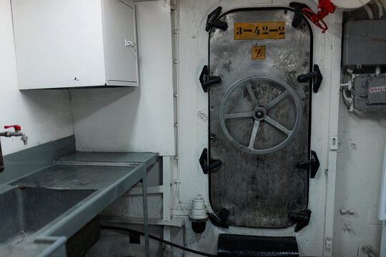 Metallic room includes bolted vault-style door, rotating wheel, and utility sink. White paint and gray steel dominate compact, functional space.Industrial ship interior with metal door and heavy wheel