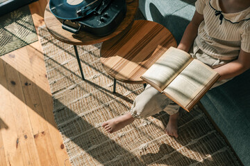 Child sitting on sofa reading book next to vinyl record player in cozy living room, sunlight...