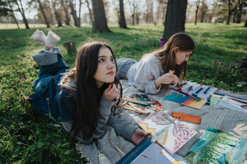 Two creative teenagers are painting masterpieces on a sunny day in a park. Lying on a blanket,...