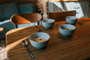 Close-up of four bowls with cereal and milk on a wooden dining table in a cozy kitchen with soft morning light