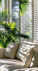 Fototapeta premium A bright sunroom with white horizontal curtains, green palm leaves and a cream-colored sofa, bathed in warm sunlight.