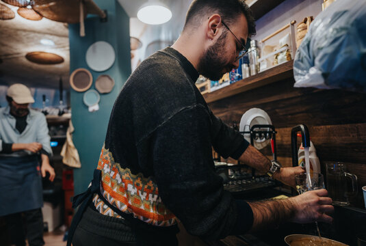 A barista washes a glass while a colleague prepares the bar area in a cozy cafe. The setting features a stylish wooden counter, warm lighting, and a charming professional atmosphere.
