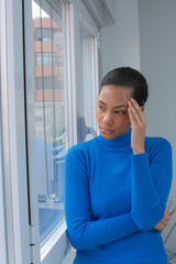 Vertical portrait worried latin young woman standing next to a window having a headache wearing...