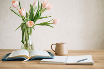 Open Bible on a wooden table and ceramic coffee mug and a vase with tulips.