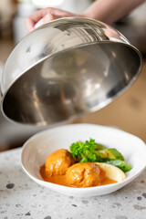 A hand lifts a metal cloche, revealing a plate with meatballs in sauce, mashed potatoes, and fresh greens on a speckled countertop. The dish is carefully plated, showcasing a gourmet meal presentation