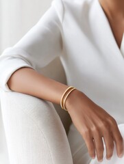 Elegant gold bangles adorn the wrist of a woman in a white blouse seated in a neutral-toned chair Minimalist jewelry and simple clothing