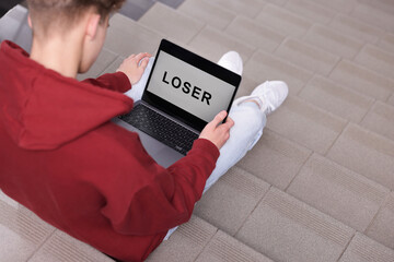 Cyber bullying. Upset teenage boy looking at laptop with text You Loser on stairs indoors, closeup