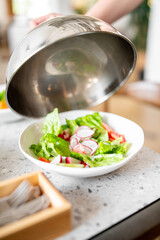A fresh salad with lettuce, radishes, and tomatoes in a white bowl is being uncovered by a person holding a stainless steel lid