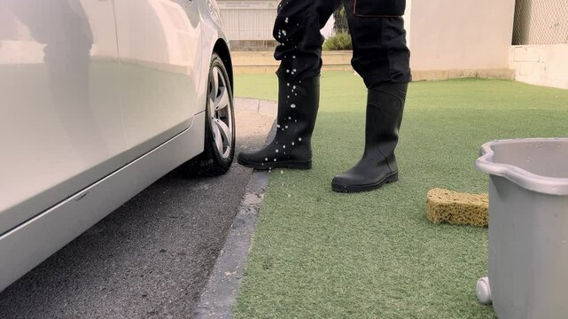Person wearing black rubber boots carefully washes a gray car, using a bucket of soapy water and a sponge, focusing on the lower panels and rocker area