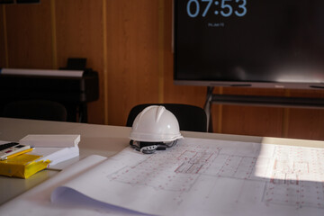 Architectural blueprints spread out on a table in a modern office, with a white safety helmet...