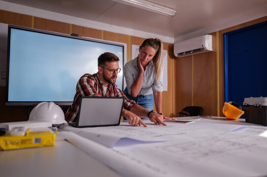 Two engineers are examining a blueprint on a table in a construction office container, utilizing a laptop for their analysis and pointing at specifics on the plan