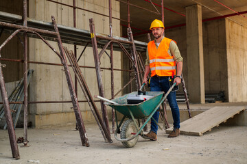 Construction worker wearing safety vest and helmet pushing wheelbarrow with tools and materials at...