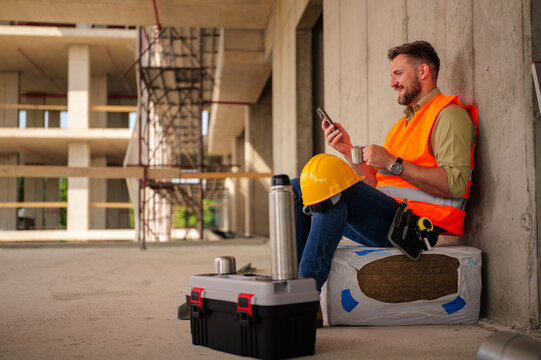 Construction worker taking a break from work, sitting and enjoying a cup of coffee while checking messages on his smartphone, with his toolbox and hardhat nearby