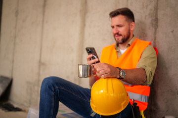 Construction worker in a safety vest enjoying a coffee break while sitting next to his helmet,...