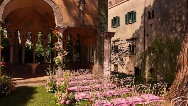 Drone view of a luxury destination wedding setup in Ravello, Italy. Pink chairs, fresh flowers at green lawn, and coastal views at Villa Cimbrone Amalfi Coast. Romantic outdoor ceremony in summer sun