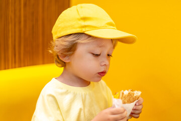 Young child wearing a yellow hat and shirt eats a cheeseburger in a vibrant restaurant, enjoying fast food meal in a casual dining environment. Kids diet, bad food choices, early junk food addiction