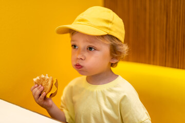 Young child wearing a yellow hat and shirt eats a cheeseburger in a vibrant restaurant, enjoying fast food meal in a casual dining environment. Kids diet, bad food choices, early junk food addiction