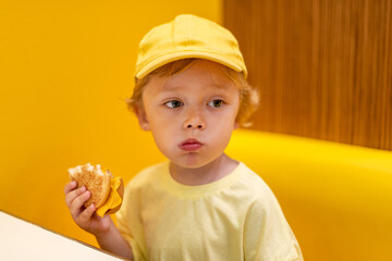 Young child wearing a yellow hat and shirt eats a cheeseburger in a vibrant restaurant, enjoying fast food meal in a casual dining environment. Kids diet, bad food choices, early junk food addiction