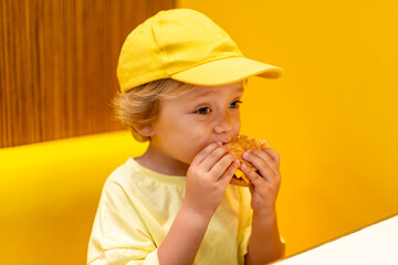 Young child wearing a yellow hat and shirt eats a cheeseburger in a vibrant restaurant, enjoying fast food meal in a casual dining environment. Kids diet, bad food choices, early junk food addiction