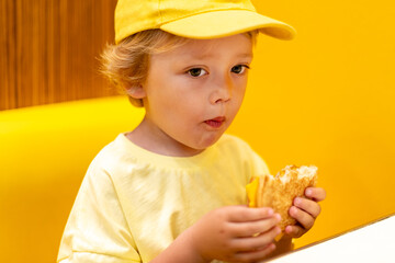 Young child wearing a yellow hat and shirt eats a cheeseburger in a vibrant restaurant, enjoying fast food meal in a casual dining environment. Kids diet, bad food choices, early junk food addiction