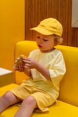 Young child wearing a yellow hat and shirt eats a cheeseburger in a vibrant restaurant, enjoying fast food meal in a casual dining environment. Kids diet, bad food choices, early junk food addiction