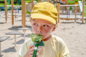 A young boy is sipping water from a bottle while resting outdoors at a sunny playground. He is wearing a bright yellow cap and enjoying his time in the warm summer weather.