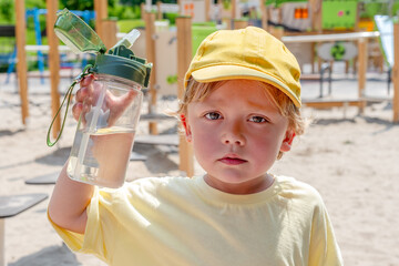 A young boy is holding a water bottle while resting outdoors at a sunny playground. He is wearing a bright yellow cap and enjoying his time in the warm summer weather. Hot summer