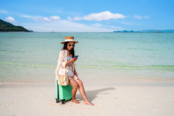 Young woman traveler with luggage using mobile phone at beautiful tropical sand beach, Summer vacation and Travel concept