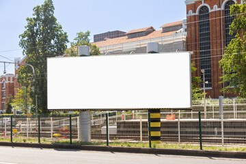 Obraz premium Blank billboard on a sunny day in an urban setting with historic brick buildings and trees in the background. Perfect for advertising mockups, marketing campaigns, or promotional designs.