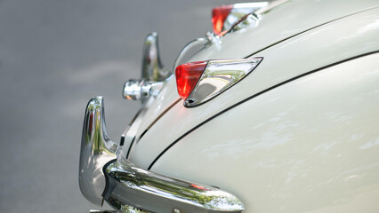Polished chrome bumper and red tail light on a classic ivory-colored vintage car, with tree reflections on a smooth rear fender.