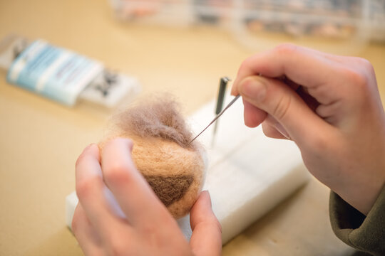 Close-up of artist hands using needle felting technique attaching small piece of wool on handmade toy figure, crafting process of creating cute animal from natural materials