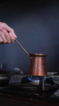 An authentic and artisanal mood is captured as a hand carefully holds a traditional hammered copper cezve pot directly over a blue gas flame