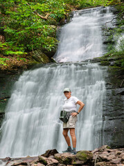 Active woman in her 60's poses in front of a waterfall after a hike through the forest.
