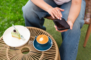 Close-up of a woman holding a cappuccino with latte art and a smartphone during a relaxing outdoors break featuring a plate with a dessert slice on a woven table.