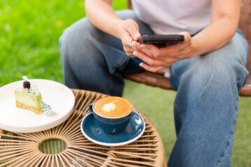 Close-up of a woman holding a cappuccino with latte art and a smartphone during a relaxing outdoors break featuring a plate with a dessert slice on a woven table.