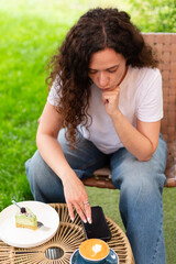 Close-up of a woman holding a cappuccino with latte art and a smartphone during a relaxing outdoors break featuring a plate with a dessert slice on a woven table.