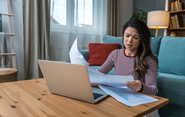 Young woman struggling with focus while working on laptop on floor, surrounded by printed analytic,...