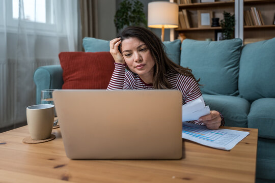 Young woman struggling with focus while working on laptop on floor, surrounded by printed analytic, battling ADHD, mental fatigue, burnout, and the pressure of modern multitasking expectations
