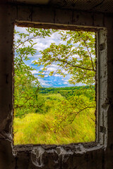 Green landscape seen through a damaged window frame of an abandoned building.
