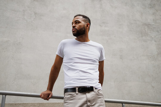 Stylish young african american man in t-shirt standing near railing and building outdoors
