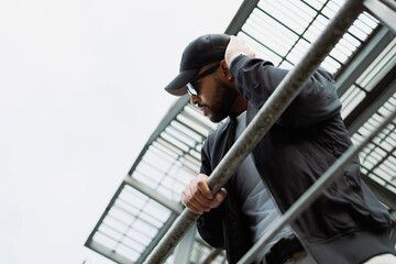 Stylish african american man in sunglasses and baseball cap standing on fire escape