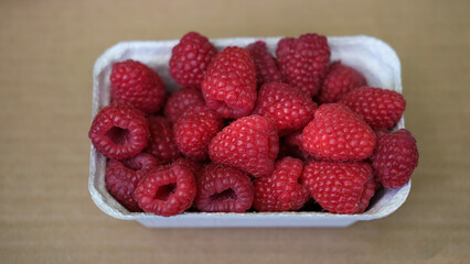 Close-up of ripe organic raspberries in a carton box on wooden surface. Fresh summer berries, natural vegan snack, local market produce.