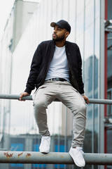 African american man in stylish bomber jacket sitting on railing on urban street