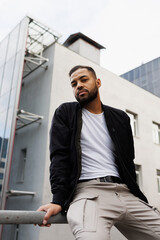 Stylish young african american man in bomber jacket sitting on railing on street